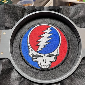 Könnte beinhalten: Gestickter Aufnäher mit Totenkopf und Blitzmotiv auf rotem und blauem Hintergrund. Der Aufnäher wird in einem grauen Stickrahmen gehalten und liegt auf einer schwarzen, strukturierten Oberfläche. Ein klassisches Emblem.