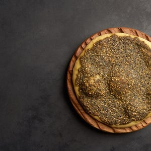 May include: A round flatbread topped with a green and black zaatar spice blend, served on a wooden plate.