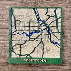 Puede incluir: Un mapa de madera de Sherbrooke, con un borde verde y letras doradas. El mapa presenta un diseño detallado de las calles, con azul representando el río y las vías fluviales. El mapa tiene forma cuadrada y se muestra sobre una superficie de madera.