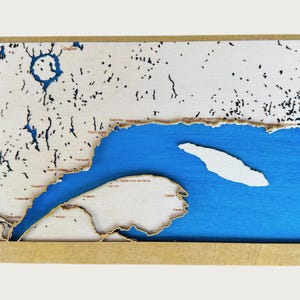 Peut inclure: Une carte en bois d'une région côtière, avec une mer bleue et une masse terrestre de couleur claire avec des accents sombres. La carte est encadrée dans un cadre en bois brun clair. Des noms de lieux sont inclus.