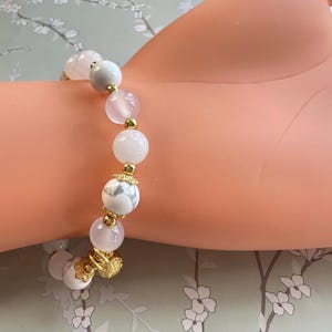 May include: A beaded bracelet featuring a mix of white, pink, and gray round stones. The bracelet includes gold-colored accents and decorative elements. The bracelet is displayed on a light-colored background.
