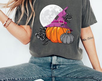 Puede incluir: Una camiseta gris con un gráfico de una calabaza con un sombrero de bruja y una luna llena en el fondo. La calabaza es naranja y el sombrero es morado con una hebilla negra. Hay rosas negras alrededor de la calabaza.