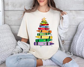 Könnte beinhalten: Ein cremefarbenes Kurzarm-T-Shirt mit einer Grafik eines Weihnachtsbaums, der aus gestapelten, bunten Wachsmalstiften besteht. Der Baum ist mit einem gelben Stern gekrönt und mit kleinen, bunten Lichterketten verziert, die um die Stifte gewickelt sind.