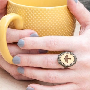 May include: A person's hand wearing a ring with a wooden bird design. The ring is set in a silver oval frame. The person is holding a yellow mug.