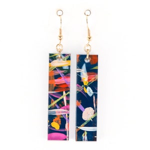 May include: A pair of dangle earrings with abstract colorful paint splatter designs on a black background. The earrings have gold hooks.