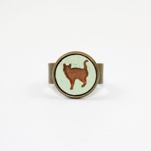 May include: A round, brass ring with a light green background and a brown silhouette of a cat.