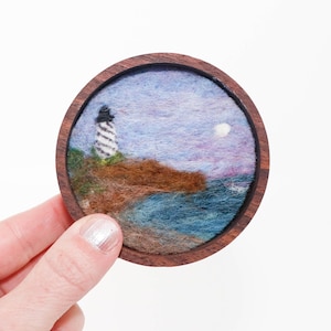 May include: A round wooden coaster featuring a felted landscape scene. The artwork depicts a lighthouse on a cliff overlooking the ocean under a purple and blue sky with a full moon. The coaster is held by a hand.