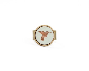 Puede incluir: Un anillo blanco con una banda de latón y una silueta de colibrí marrón en la parte superior.