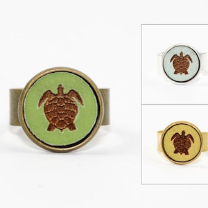 Peut inclure: Ensemble de trois bagues réglables avec un motif de tortue marron sur fond vert, bleu et jaune. Les bagues sont en métal et ont une forme ronde.