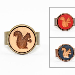 May include: Three rings with a wooden squirrel silhouette design. The rings are made of metal and have a round, flat top. The squirrel silhouette is brown and is set against a beige, red, and blue background.