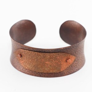 May include: A wide copper cuff bracelet with a hammered texture and a smaller copper plate inset in the front. The plate has a hammered texture and is slightly oxidized.