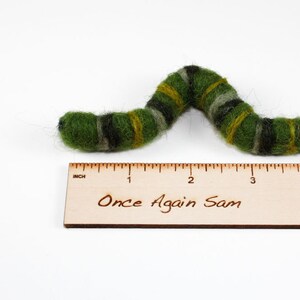 Pu&ograve; includere: Un giocattolo a forma di bruco in feltro verde, giallo e nero che misura circa 10 cm di lunghezza. Il giocattolo &egrave; su un righello di legno con le parole "Once Again Sam" scritte su di esso.