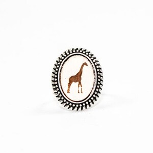 May include: Oval-shaped ring with a silver-colored band and a detailed border. The ring features a brown giraffe silhouette on a white background. The giraffe is standing upright, with its long neck and legs visible.