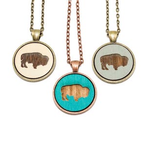 May include: Three necklaces with round wooden pendants featuring a silhouette of a bison. The pendants are in beige, gray, and turquoise.