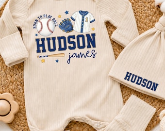 Peut inclure: Un ensemble composé d'une grenouillère à manches longues et d'un bonnet en tissu côtelé beige. La grenouillère présente un motif de baseball avec une balle, un gant, un maillot, une batte et le texte "BORN TO PLAY BALL", "HUDSON", "james".