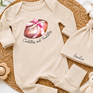 May include: A beige ribbed long-sleeve romper and matching beanie set. The romper features a graphic of a brown football wrapped in a pink ribbon with the text Cuddles and Huddles. The beanie is personalised with the name Amelia in black script.