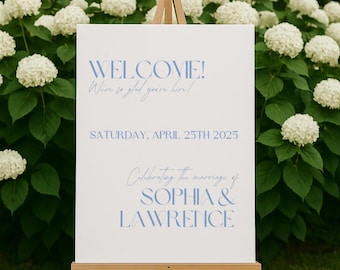 Op de afbeelding: Wit welkomsbord met blauwe tekst. Het bord zegt "WELCOME! We're so glad you're here!" en "SATURDAY, APRIL 25TH 2025" en "Celebrating the marriage of SOPHIA & LAWRENCE".