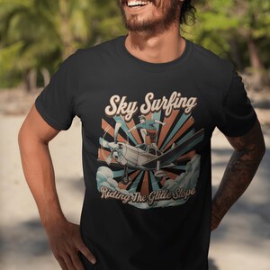 May include: A black short-sleeved t-shirt featuring a graphic print of a person standing on top of a small aeroplane in flight. The design includes the text "Sky Surfing" and "Riding The Glide Slope" with a sunburst background in blue and orange.