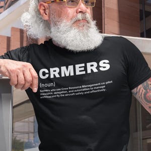 May include: Black t-shirt with the word "CRMERS" in white block letters. Below the word is the definition of CRMERS, which is "Simmers who use Crew Resource Management co-pilot assistance, delegation, and automation to manage workload and fly the aircraft safely and effectively."
