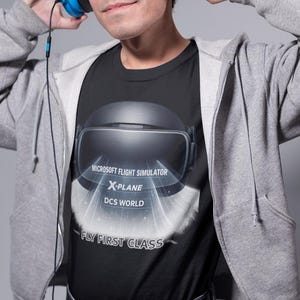 May include: A black t-shirt featuring a graphic of a virtual reality headset with light rays emanating from the lenses. The text reads MICROSOFT FLIGHT SIMULATOR, X-PLANE, DCS WORLD, and FLY FIRST CLASS. The shirt is worn under a gray zip-up hoodie.