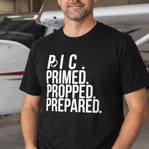 May include: Black t-shirt with white text that reads "PIC. PRIMED. PROPPED. PREPARED." The shirt is short-sleeved and is being worn by a person. A small airplane is visible in the background.
