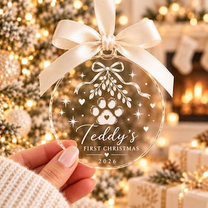 May include: A clear circular acrylic ornament featuring a white etched design of a paw print with a heart, a bow, and stars. The text reads "Teddy's FIRST CHRISTMAS" and "2026" with a small heart. A cream-colored satin ribbon is tied at the top.