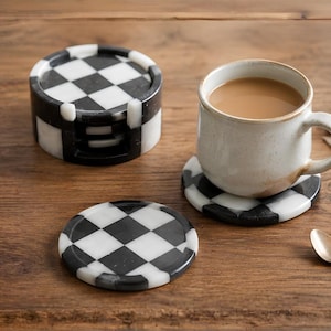 May include: A set of round stone coasters featuring a black and white checkered pattern. One coaster is placed under a ceramic mug filled with a light brown beverage, while another sits flat nearby, and a stack of coasters rests in a matching holder.
