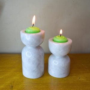 May include: Two white marble candle holders with green candles. The candle holders are hourglass shaped and have a smooth, polished finish.