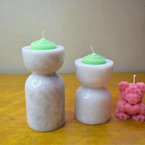 May include: Three candles in different shapes and sizes. Two candles are in white marble candle holders with green candles on top. The third candle is pink and shaped like a teddy bear.