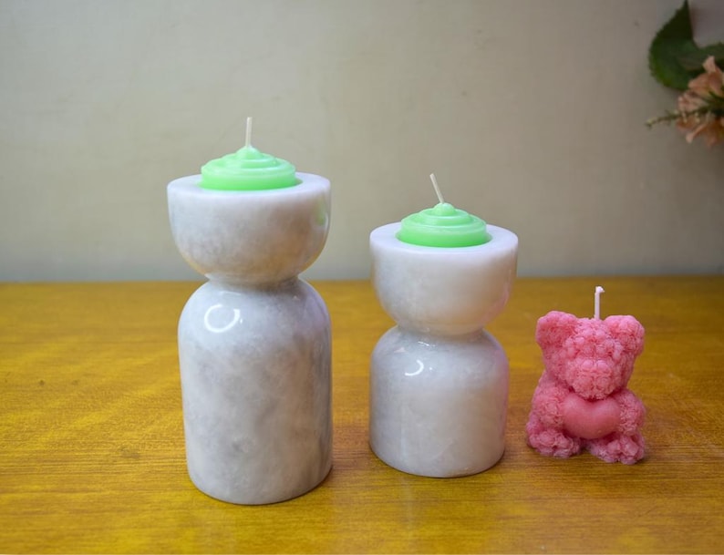 May include: Three candles in different shapes and sizes. Two candles are in white marble candle holders with green candles on top. The third candle is pink and shaped like a teddy bear.