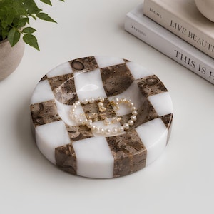May include: A circular stone tray featuring a checkered pattern of alternating white and brown marble squares. The tray has a shallow, concave center currently holding a delicate string of white pearls and small gold-toned jewelry pieces.