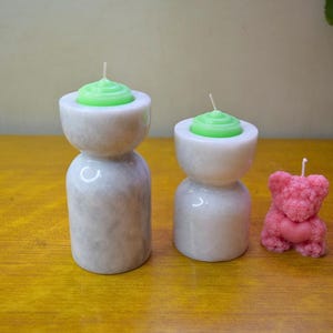 May include: Two white marble candle holders with green candles and a pink teddy bear candle.