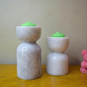 May include: Two white marble candle holders with green candles on top. A pink teddy bear candle is on the right side of the image.