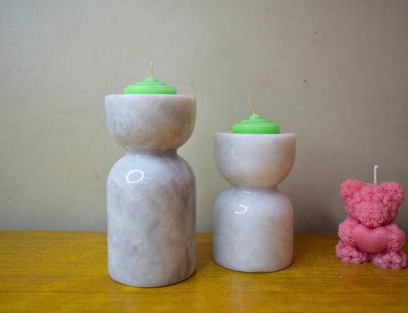 May include: Two white marble candle holders with green candles on top. A pink teddy bear candle is on the right side of the image.