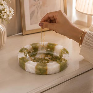 May include: A round, shallow tray featuring a checkered pattern of translucent white and olive green stone segments. The tray contains a gold-toned chain necklace with a small star-shaped pendant and a matching gold-toned ring resting inside.
