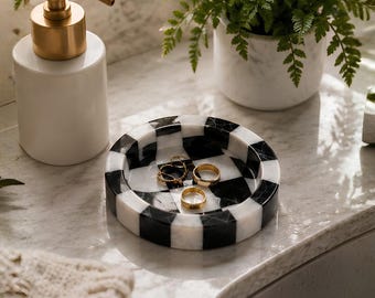 May include: A round marble trinket tray featuring a black and white checkered pattern. The tray contains two gold-toned metal rings and a delicate gold chain. The surface of the tray has a polished finish with visible natural stone veining.