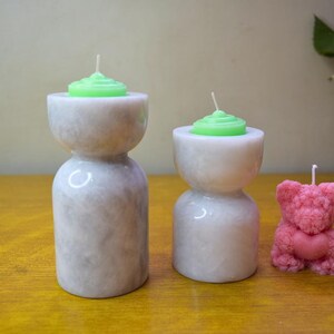 May include: Three candle holders made of white marble with a textured surface. Two of the holders have green candles and the third holder has a pink bear-shaped candle.