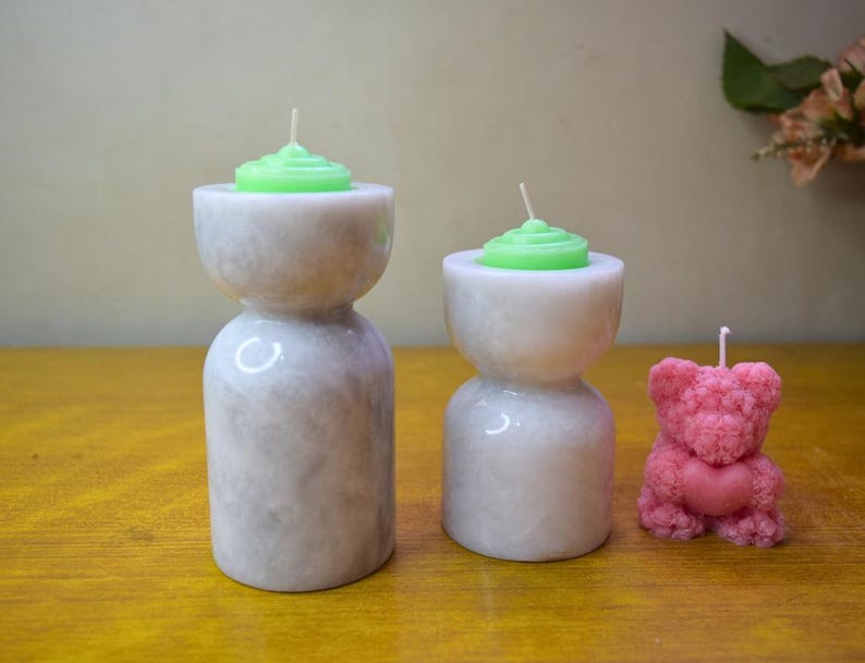 May include: Three candle holders made of white marble with a textured surface. Two of the holders have green candles and the third holder has a pink bear-shaped candle.