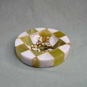 May include: A white and green marble trinket dish with a checkered pattern. The dish is filled with a gold and pearl necklace.