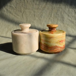 May include: Two small, cylindrical stone jars with lids. One is a creamy white color, and the other is a mix of yellow, orange, and brown. The jars have a smooth, polished finish and are likely used for storage or decoration.