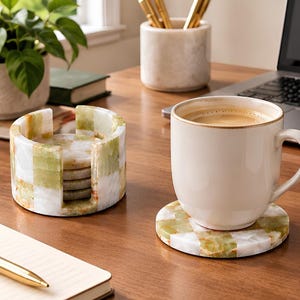 May include: A set of round stone coasters featuring a checkered pattern in shades of white and light green. The set includes a matching cylindrical holder for storage, and one coaster is placed under a white ceramic mug filled with coffee.