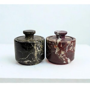 May include: Two small, round marble jars with lids. One jar is black with gold veining, and the other is burgundy with white veining. Both jars have a small, round knob on top of the lid.