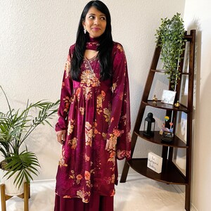 May include: A burgundy floral print salwar kameez set. The long-sleeved tunic features a V-neckline and is paired with matching wide-leg pants and a scarf. The floral pattern includes shades of peach and yellow.