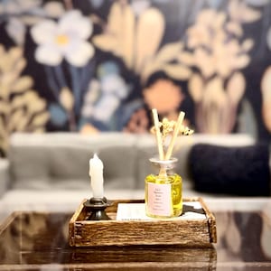 May include: A wooden tray holds a candle in a black holder, a glass bottle with reeds, and a small notepad. The bottle contains a yellow liquid and a pink label. The background features a floral wallpaper.