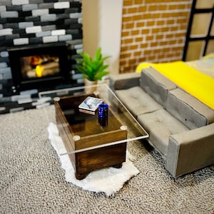 May include: A miniature living room scene featuring a dark wood coffee table with a clear glass top, a gray tufted sofa with a yellow throw blanket, a small blue cup, and a tiny book, all set on a textured gray carpet before a fireplace.