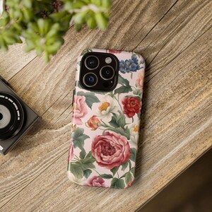 May include: A pink floral phone case with a white background and a camera lens cutout. The case features a variety of pink, red, and white flowers with green leaves.