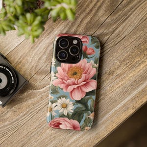 May include: A phone case with a floral design. The case features pink, white, and green flowers on a light blue background. The phone case has a camera cutout for a triple lens camera.