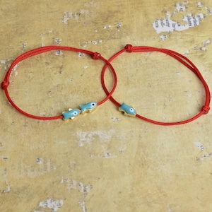 May include: Two red string bracelets, each adorned with small, turquoise fish charms with gold accents. The bracelets are knotted and designed to be adjustable. The background is a textured, tan surface.