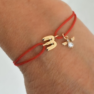 May include: Red string bracelet with gold number 3 charm and a small angel charm with a clear gemstone. The bracelet is worn on a wrist, showcasing the jewelry's design and color contrast.