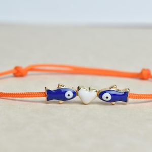 May include: An orange cord bracelet featuring three charms: two blue fish with gold accents and a white heart in the center. The bracelet is knotted at each end.